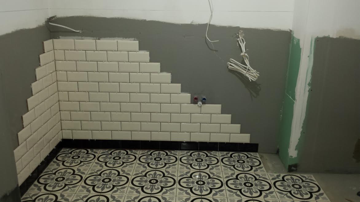 Świeżo ułożone, białe, prostokątne płytki na ścianie w łazience, ułożone schodkowo, z czarnym pasem płytek u dołu i dekoracyjną podłogą w kwiaty. Widoczne wystające rurki instalacji wodnej...