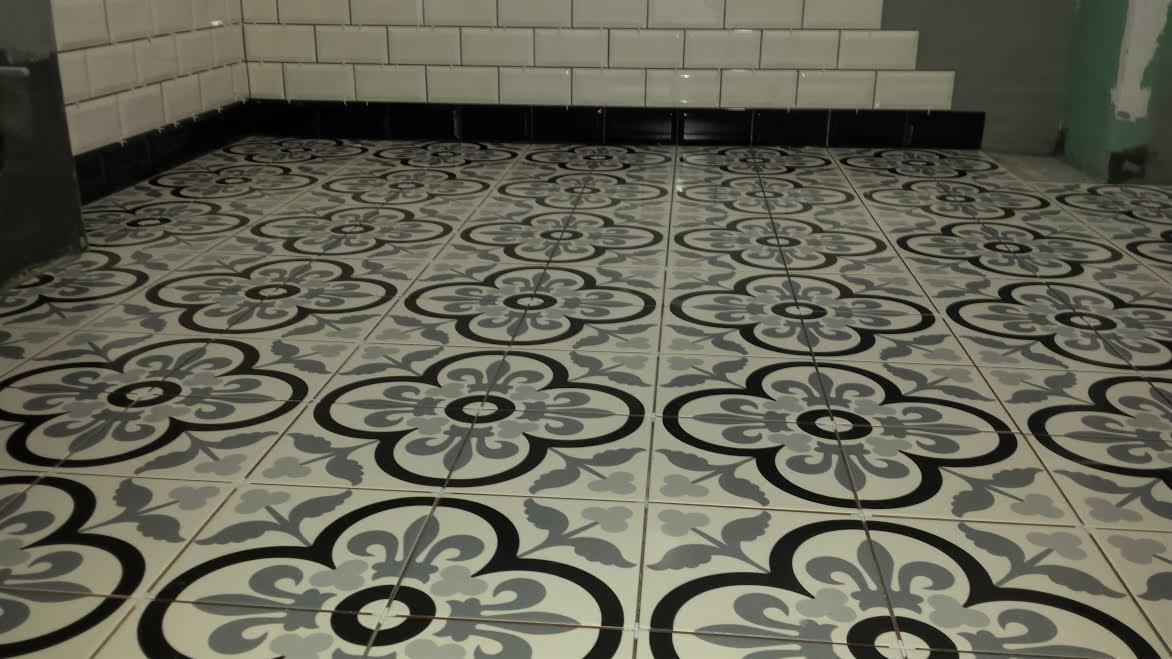 Wykończona podłoga w łazience z szarymi płytkami w kwiatowe wzory, białe płytki ścienne z czarnym pasem na dole.