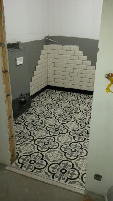 Łazienka w trakcie remontu: podłoga wyłożona wzorzystymi płytkami w odcieniach szarości i czerni, ściany częściowo pokryte białymi, prostokątnymi płytkami ułożonymi schodkowo, widoczne przygotowane...