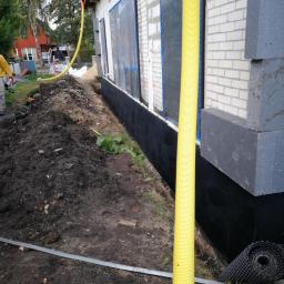 FHU Sąsiedzi Marcin Wasiluk - Przygotowanie ściany zewnętrznej budynku z białej cegły, zabezpieczonej folią okienną i czarną izolacją fundamentów, widoczna żółta rura wentylacyjna, rolka membrany kubełkowej i stos szarych...