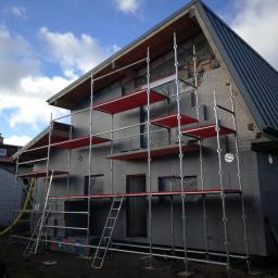 FHU Sąsiedzi Marcin Wasiluk - Dom w trakcie ocieplania, widoczne szare płyty izolacyjne na elewacji, rusztowanie z czerwonymi podestami, częściowo odsłonięty mur z cegły na szczycie, dach z blachy, niebieskie niebo z chmurami.