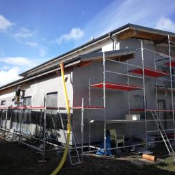 FHU Sąsiedzi Marcin Wasiluk - Budynek w trakcie ocieplania, widoczne szare bloczki, rusztowania z czerwonymi podestami, pracownik na rusztowaniu oraz żółty wąż do podawania materiału.