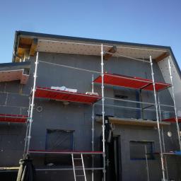 FHU Sąsiedzi Marcin Wasiluk - Dom w trakcie ocieplania styropianem, widoczne rusztowania z czerwonymi podestami, zabezpieczone okna folią, fragment dachu z widoczną izolacją.