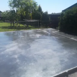 Świeżo wylana, gładka, betonowa wylewka tarasu lub pod altanę, z widocznym odpływem liniowym i śladami wilgoci, na tle zielonego ogrodu z drzewami i metalowym ogrodzeniem.