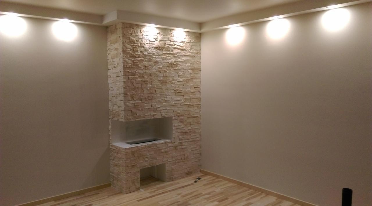 Wykończony fragment ściany w salonie z dekoracyjnym kamieniem, imitującym kominek z wnęką, oświetlony punktowymi lampami sufitowymi, na podłodze panele.