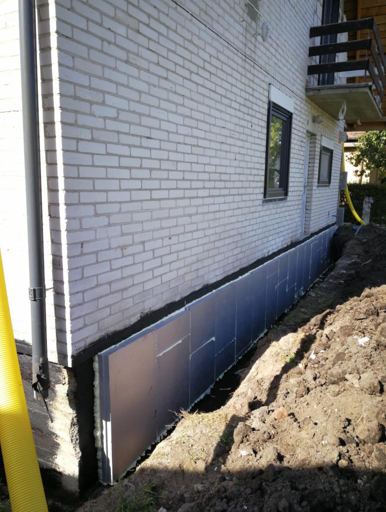 Ocieplenie fundamentów budynku z białej cegły, widoczne płyty izolacyjne ze srebrną folią, żółta rura wentylacyjna przy ścianie.