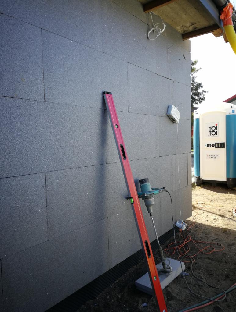 Szare płyty izolacyjne elewacji w trakcie montażu, poziomica pionowa, wiertarka z mieszadłem na betonowej płycie, przenośna toaleta w tle.