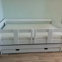 Białe, drewniane łóżko dziecięce z barierkami i wysuwaną szufladą na pościel, ustawione na panelach podłogowych, na tle jasnej, zielonej ściany.