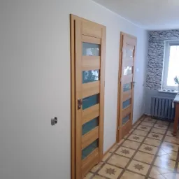 Dwa drewniane drzwi wewnętrzne z przeszkleniami, widoczne z perspektywy korytarza z wzorzystą podłogą i grzejnikiem pod oknem z dekoracyjną tapetą.