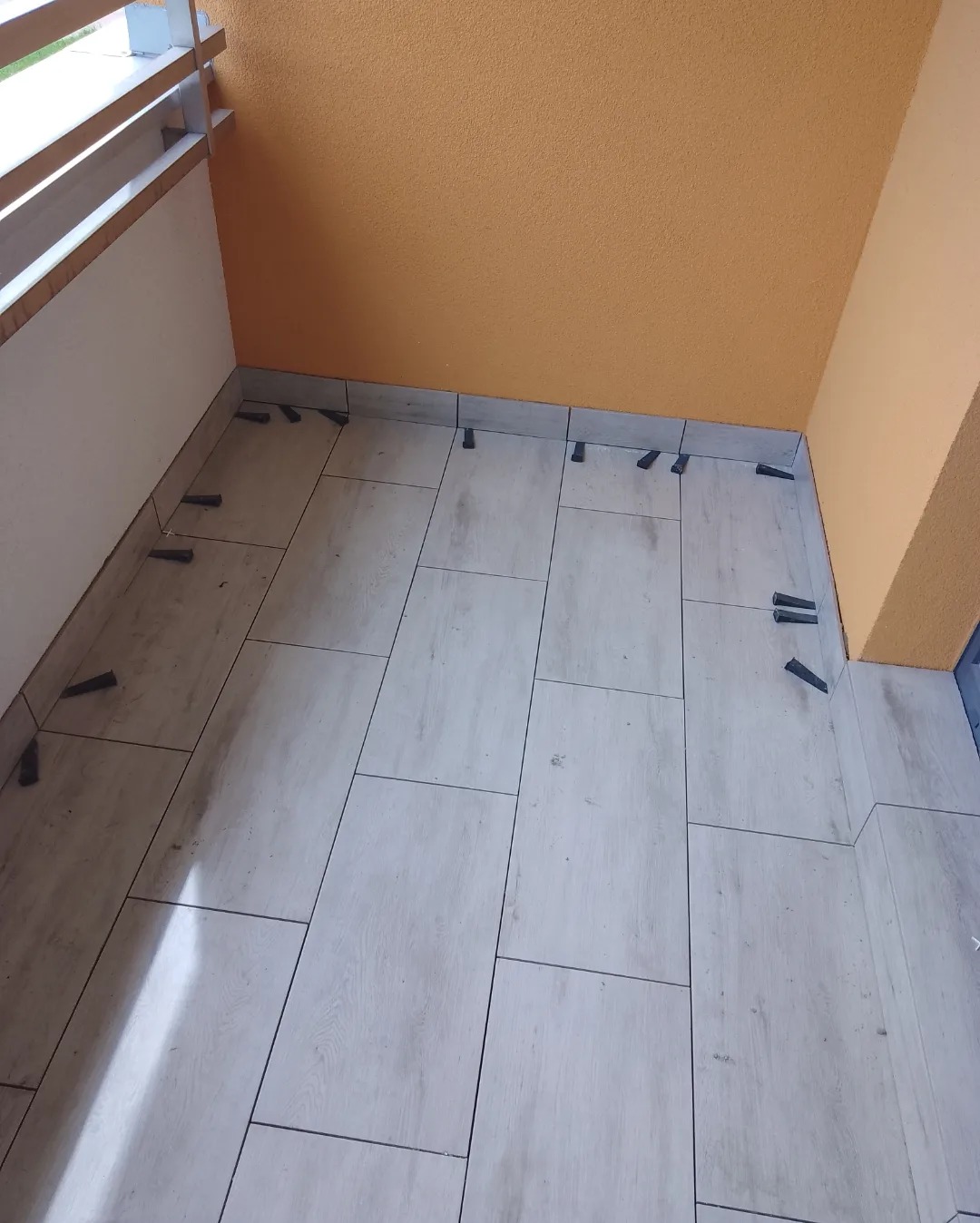 Balkon w trakcie układania jasnych, podłużnych płytek podłogowych z użyciem czarnych klinów dystansowych, pomarańczowa ściana w tle.