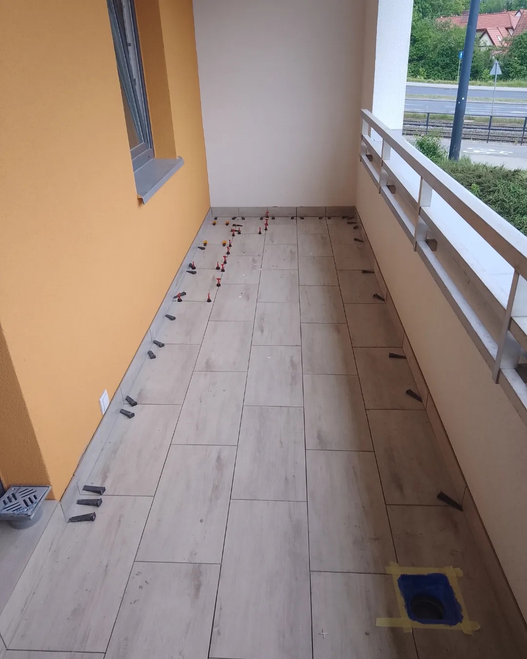 Balkon w trakcie układania płytek podłogowych; widoczne płytki w odcieniach beżu i brązu, system poziomowania płytek z czerwonymi klinami i czarnymi klipsami, oraz zabezpieczony odpływ liniowy.
