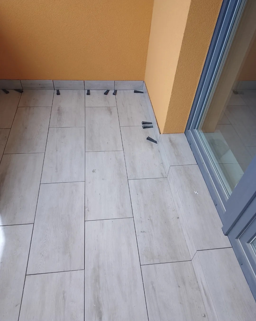 Układanie jasnych, drewnopodobnych płytek na balkonie z użyciem klinów poziomujących; widoczna pomarańczowa ściana i rama okienna.