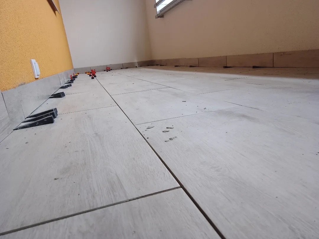 Układanie jasnych, podłużnych płytek podłogowych z użyciem klinów poziomujących, widoczne fragmenty ścian w odcieniach żółci i beżu.