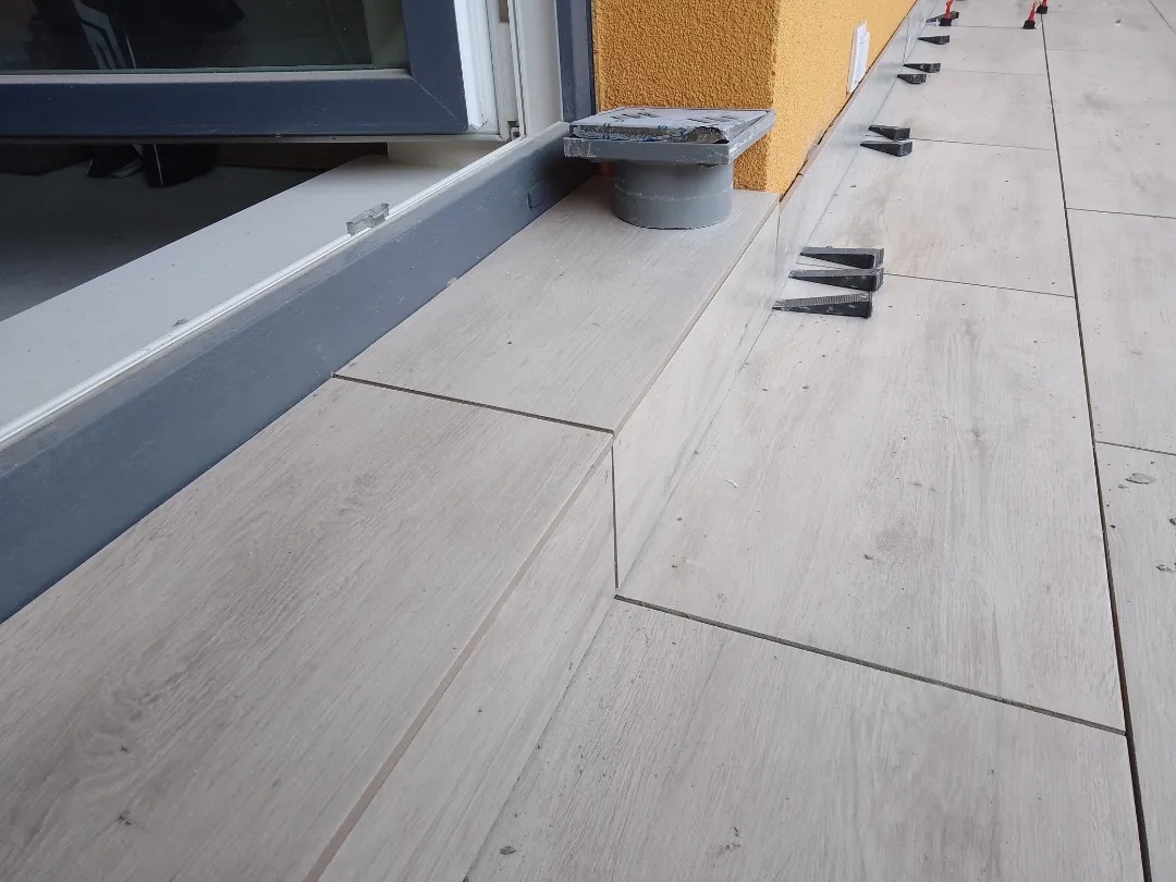 Balkon wyłożony płytkami imitującymi drewno, widoczne kliny poziomujące, odpływ liniowy przy ścianie budynku.