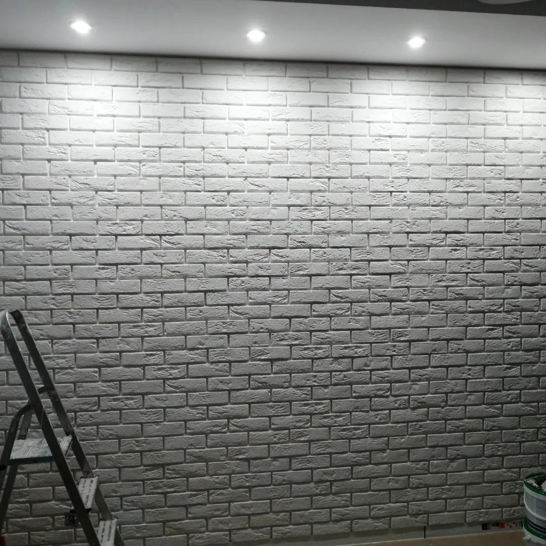 Ściana wykończona białą cegłą dekoracyjną z widoczną drabiną malarską i wiadrem z farbą w rogu, oświetlona punktowymi lampami sufitowymi.