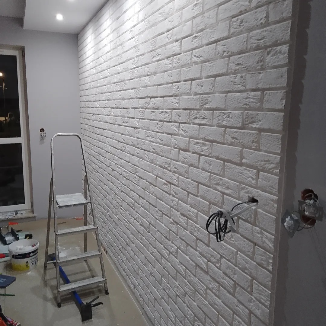 Ściana wykończona białą cegłą dekoracyjną z widoczną instalacją elektryczną, drabina malarska i akcesoria malarskie na podłodze, w trakcie prac wykończeniowych.