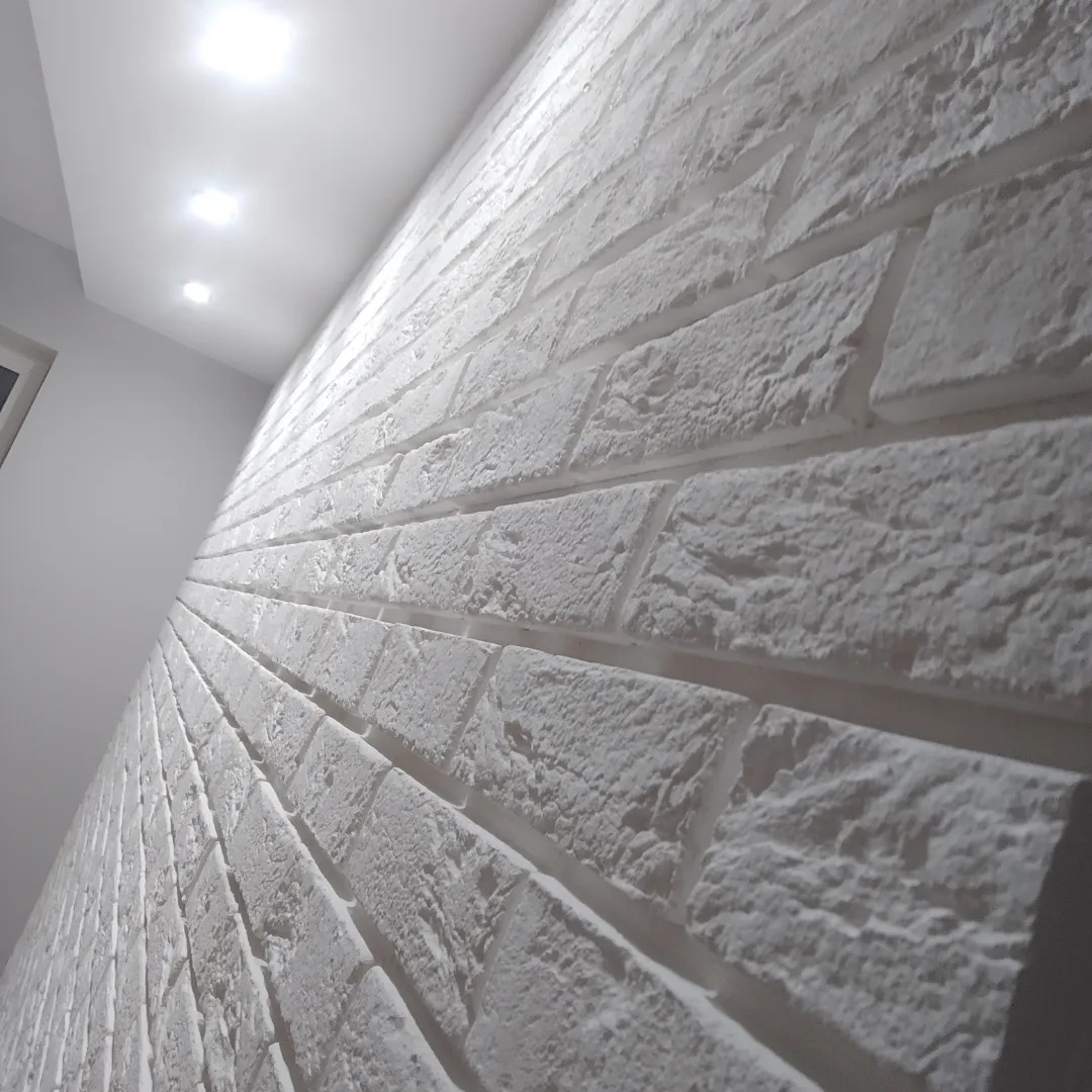 Ściana wykończona białymi, dekoracyjnymi cegłami z widoczną fakturą, oświetlona przez punktowe lampy sufitowe, z perspektywą skierowaną wzdłuż ściany.