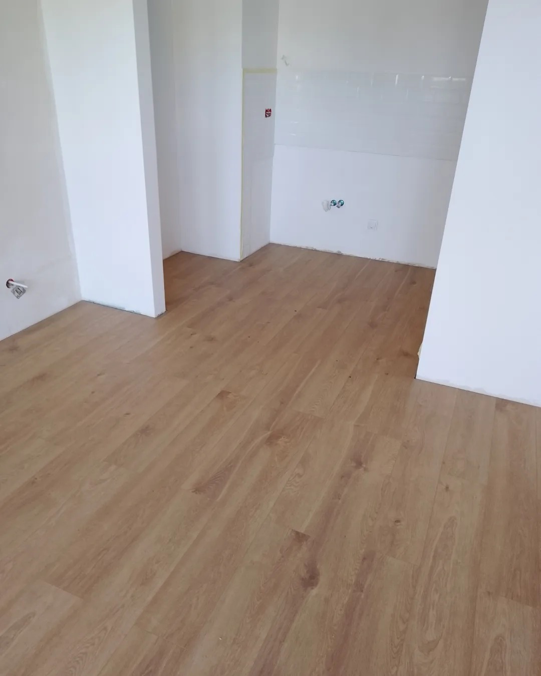 Jasne panele podłogowe ułożone w pustym pomieszczeniu z białymi ścianami i fragmentem białej ściany wyłożonej płytkami, widoczne gniazdka elektryczne.