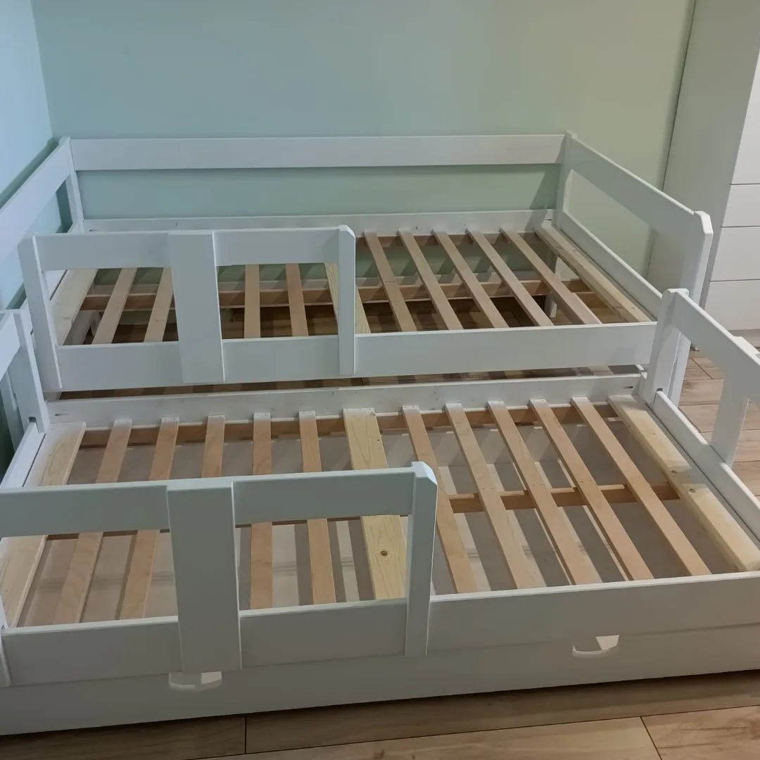 Białe, drewniane łóżko wysuwane z barierką ochronną, widoczne szczebelki stelaża, na podłodze panele w odcieniach brązu, w tle ściana w kolorze miętowym.