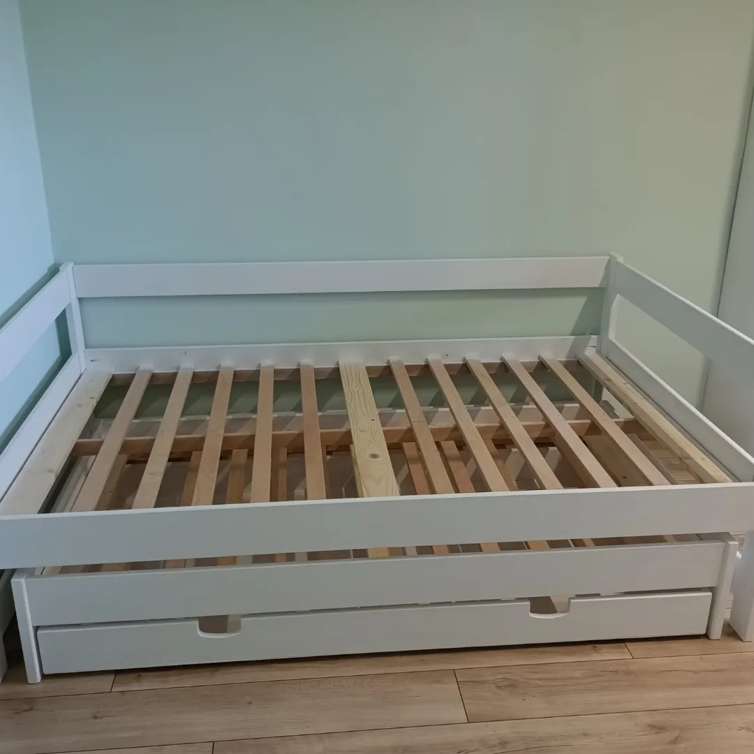 Biała, drewniana rama łóżka z wysuwaną szufladą na drewnianej podłodze, widoczne listwy pod materac, na tle jasnej ściany.