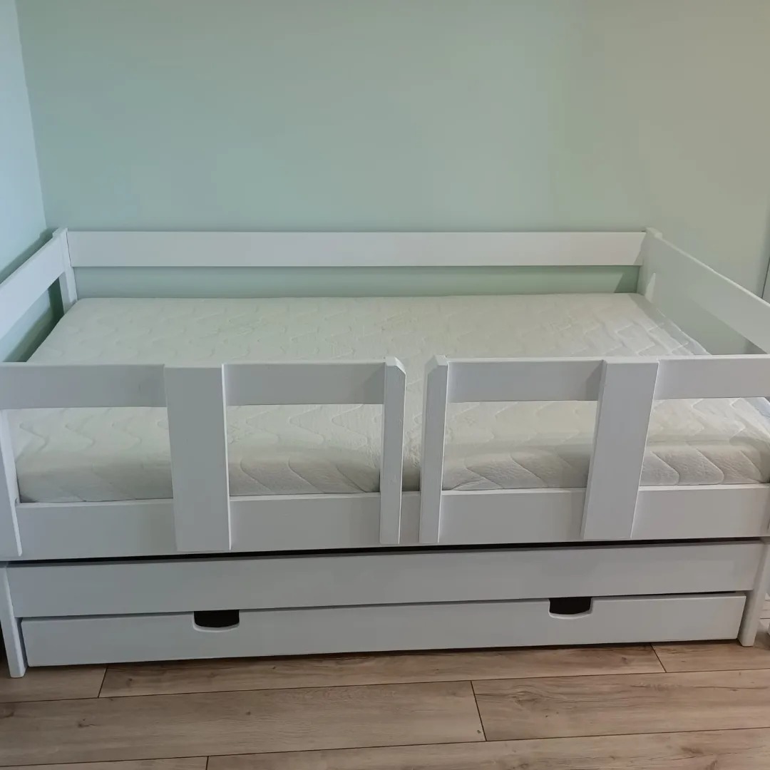 Białe, drewniane łóżko dziecięce z barierkami i wysuwaną szufladą na pościel, ustawione na panelach podłogowych, na tle jasnej, zielonej ściany.