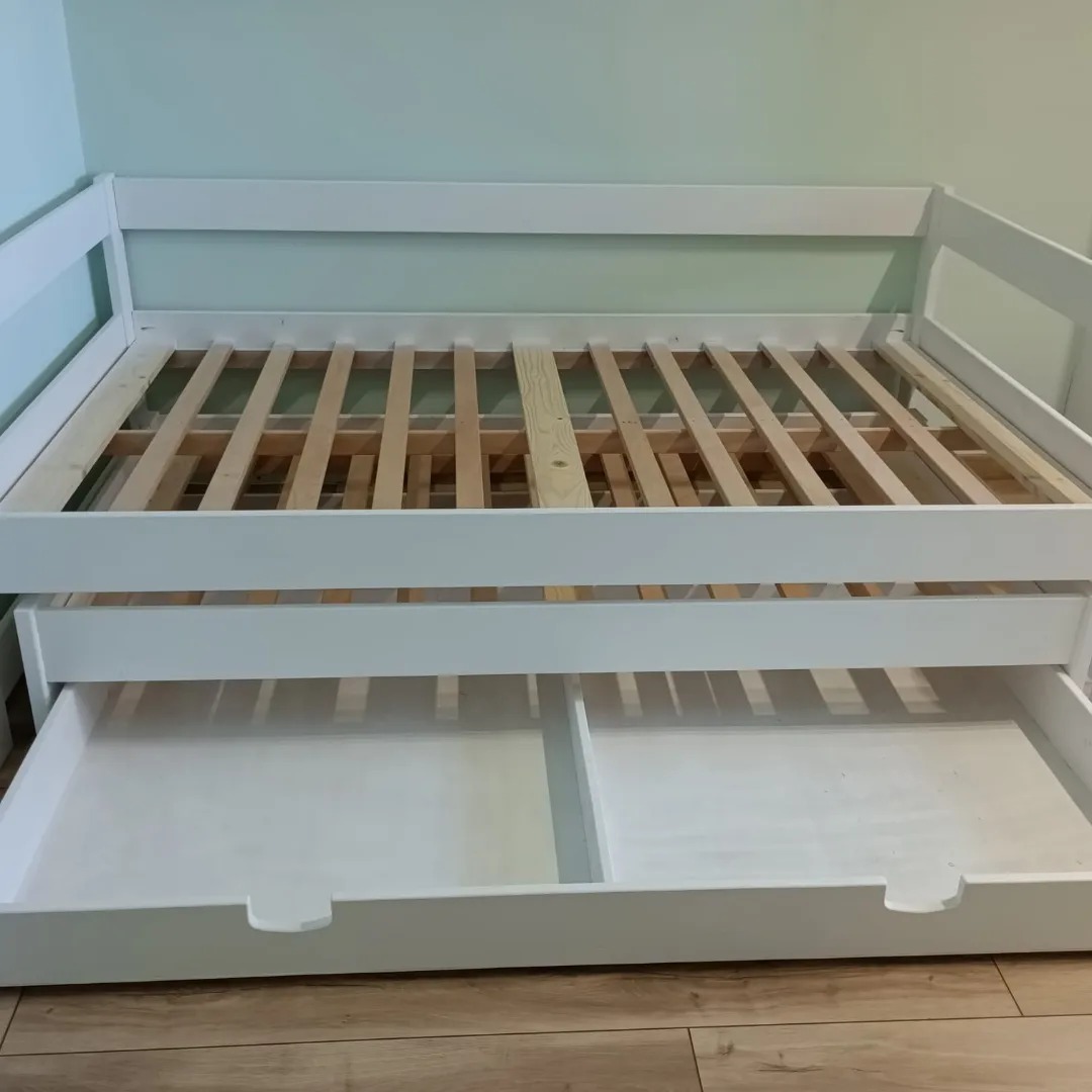 Białe drewniane łóżko z wysuwaną szufladą i drewnianym stelażem, ustawione na podłodze w panelach, na tle jasnej ściany.