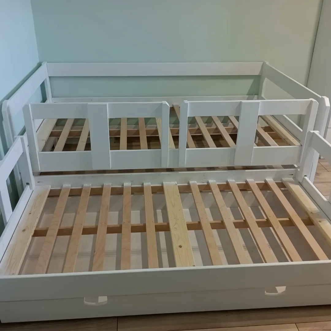 Białe, drewniane łóżko wysuwane z barierkami, widoczne drewniane stelaże i szuflada w dolnej części, na tle jasnej ściany.