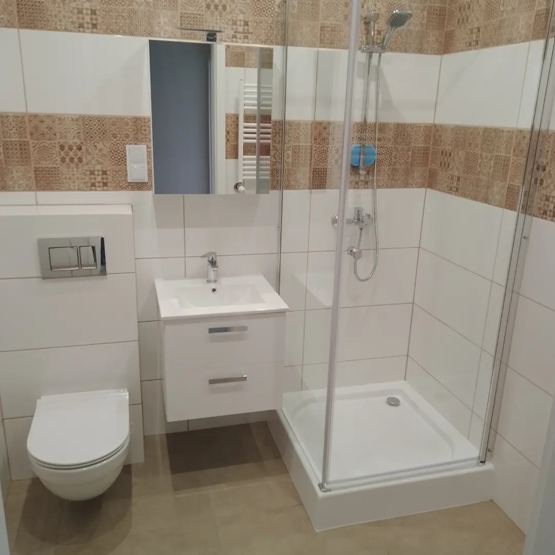 Nowoczesna łazienka z kabiną prysznicową, umywalką z szafką, toaletą i lustrem. Białe płytki kontrastują z pasem płytek dekoracyjnych w odcieniach brązu. Widoczny zestaw podtynkowy WC.