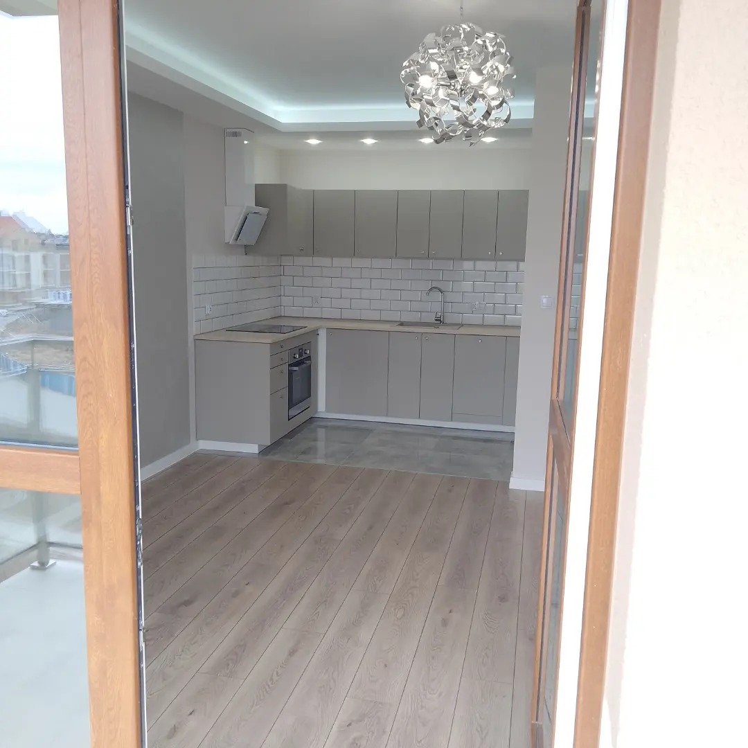 Widok na nowocześnie urządzoną kuchnię w odcieniach szarości z drewnianą podłogą, widzianą przez otwarte drzwi balkonowe; w tle widoczne zabudowania miejskie.