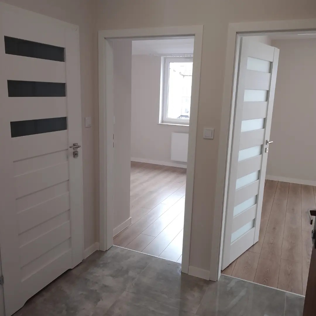 Wnętrze mieszkania po remoncie: widok na dwa białe, nowoczesne drzwi z przeszkleniami, jeden zamknięty, drugi otwarty, z widokiem na parkiet i okno w tle.