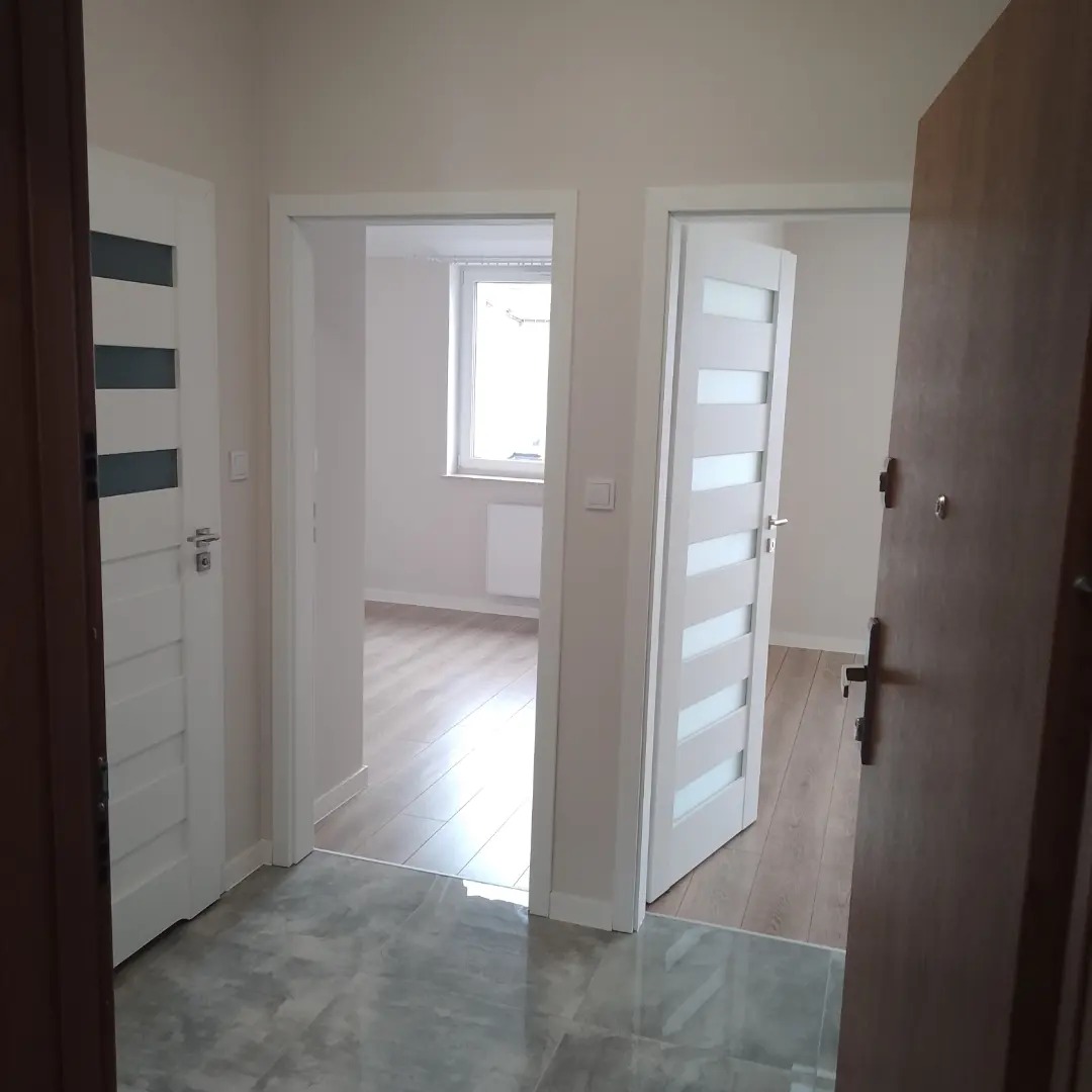 Wnętrze mieszkania po remoncie: widok na trzy drzwi wewnętrzne z różnymi wzorami, szara, błyszcząca podłoga w korytarzu, jasne ściany i drewniana podłoga w pokojach w tle.