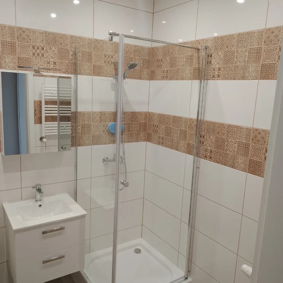 Wykończona łazienka z kabiną prysznicową ze srebrnymi profilami, białą umywalką z szafką, lustrem i poziomym pasem dekoracyjnych płytek w kolorze beżowym.