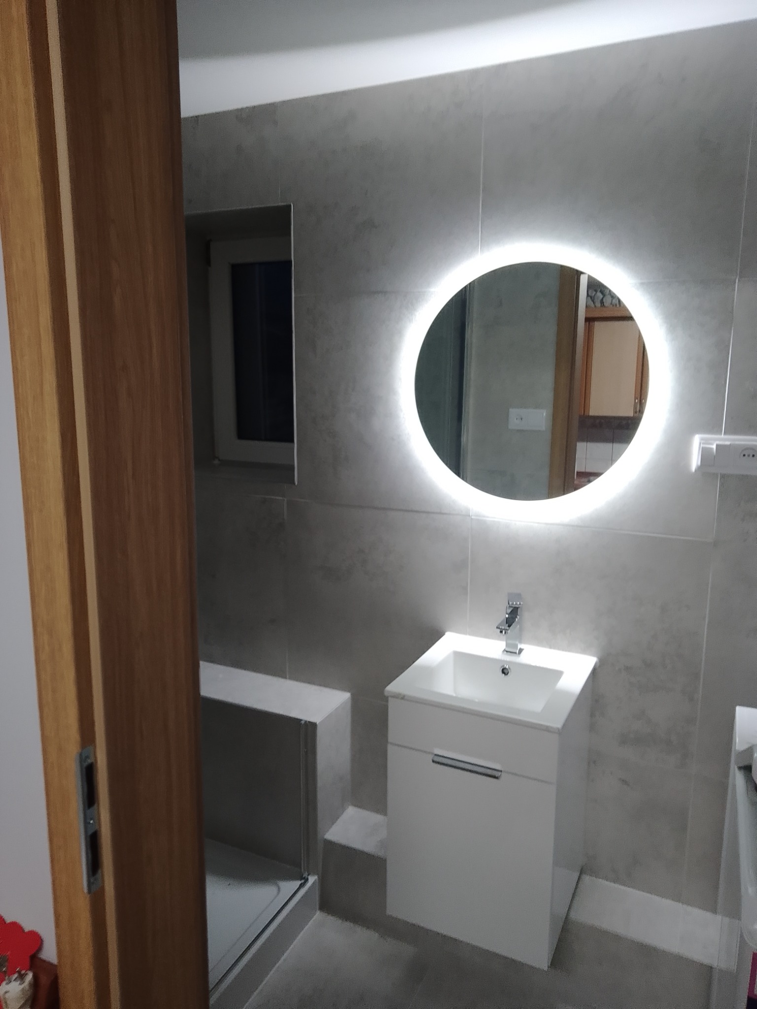 Nowoczesna łazienka z szarą płytką, okrągłym lustrem z podświetleniem LED nad białą szafką z umywalką i widocznym fragmentem kabiny prysznicowej za drewnianymi drzwiami.