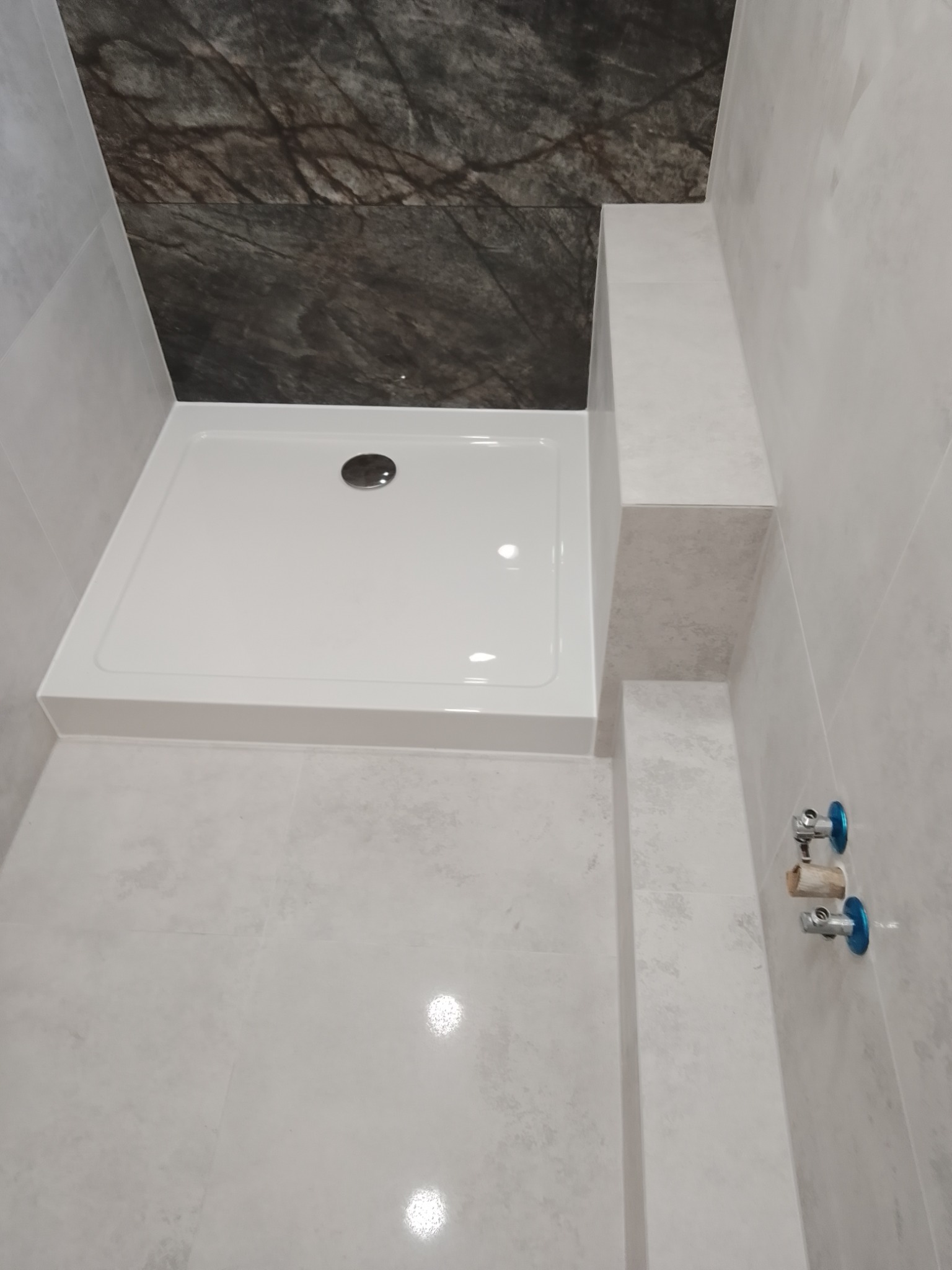 Nowoczesny brodzik z odpływem, otoczony jasnymi płytkami imitującymi kamień, z ciemnym akcentem na górze i widocznymi zaworami wodnymi na ścianie.