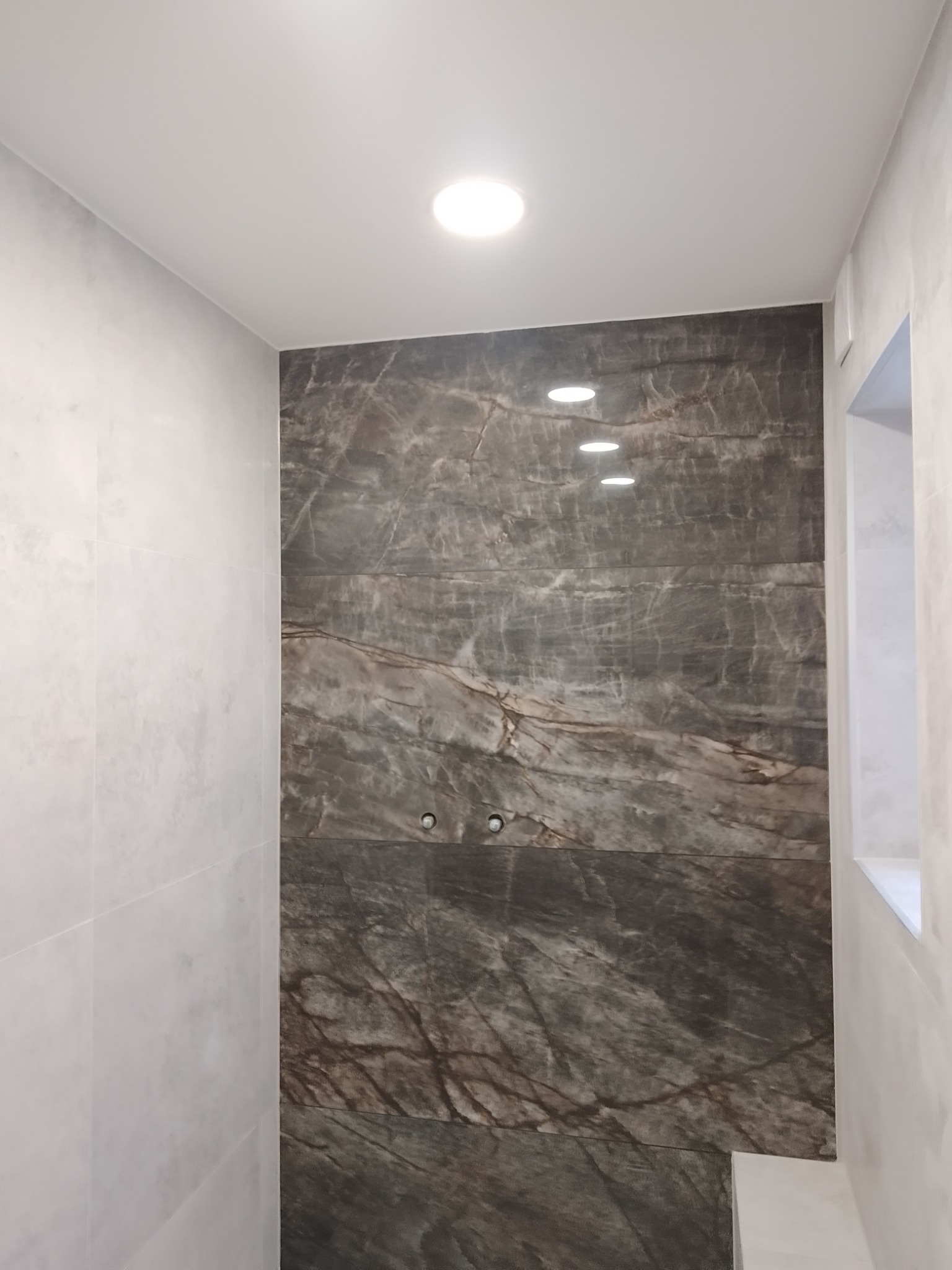 Wykończona łazienka z dużymi płytami imitującymi marmur na ścianie prysznicowej, jasnymi płytkami na pozostałych ścianach i wpuszczonym oświetleniem punktowym.