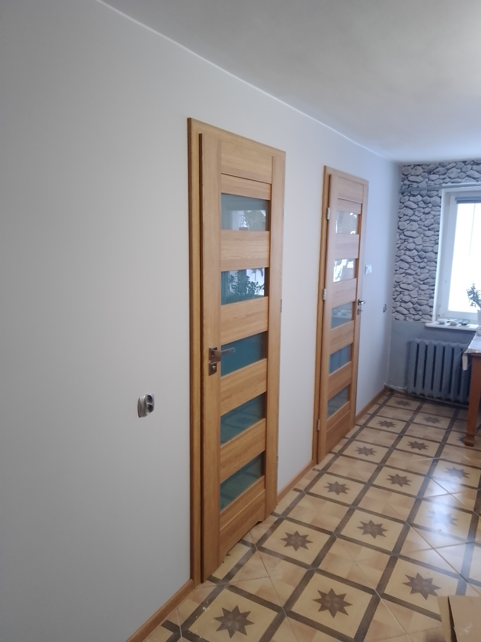 Dwa drewniane drzwi wewnętrzne z przeszkleniami, widoczne z perspektywy korytarza z wzorzystą podłogą i grzejnikiem pod oknem z dekoracyjną tapetą.