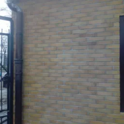 Ściana budynku z żółtej cegły z brązową rynną i fragmentem okna, widoczne ogrodzenie i drzewa w tle.