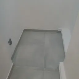 Wykończone wnętrze z szarymi płytkami podłogowymi i białymi ścianami z szarą listwą przypodłogową, widoczny włącznik światła po lewej stronie, ujęcie z góry.