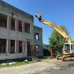 Żółta koparka Liebherr z uniesionym ramieniem i łyżką na tle częściowo rozebranego, dwupiętrowego budynku z pustymi otworami okiennymi, widoczny znak ostrzegawczy i gruz.