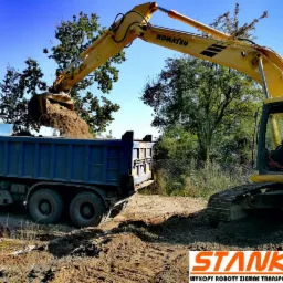 Żółta koparka Komatsu z operatorem w kabinie, ładuje ziemię do niebieskiej ciężarówki na tle drzew.