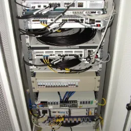 Szafa rack z zamontowanymi urządzeniami sieciowymi, kablami światłowodowymi i elektrycznymi, widoczne patch panele, switche i listwy zasilające, ułożone w uporządkowany sposób.