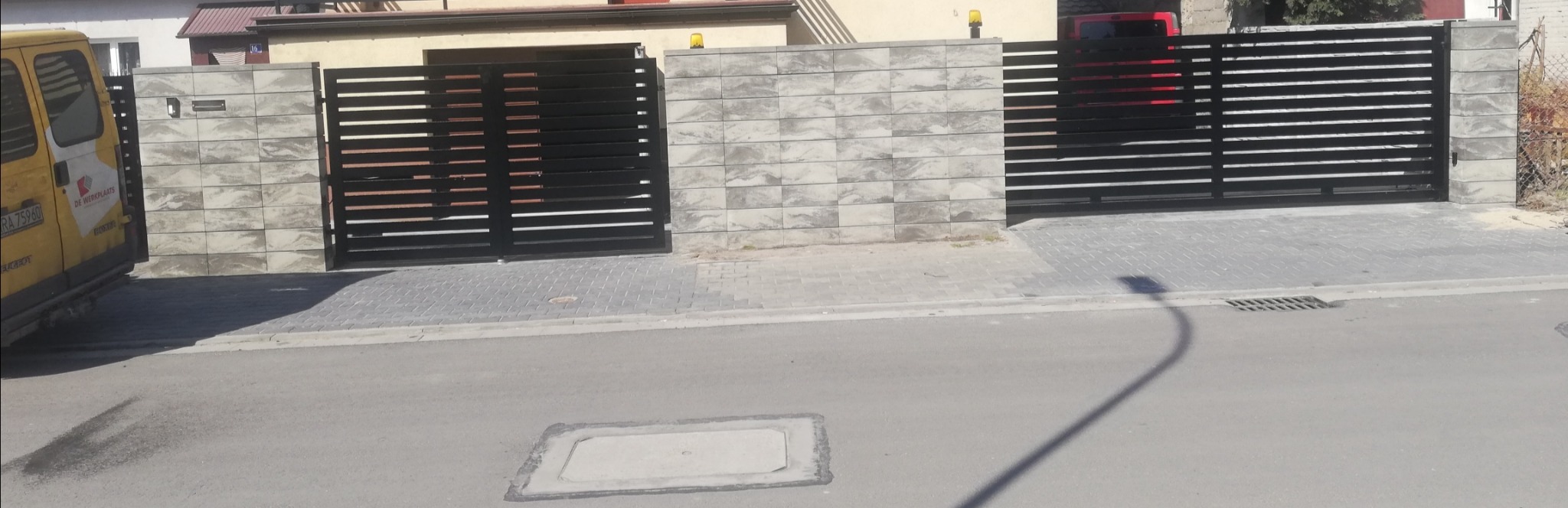 Nowoczesna, pozioma brama metalowa z czarnymi panelami i betonowymi słupkami w kolorze szarym, z widocznym żółtym samochodem dostawczym i fragmentem czerwonego auta w tle.