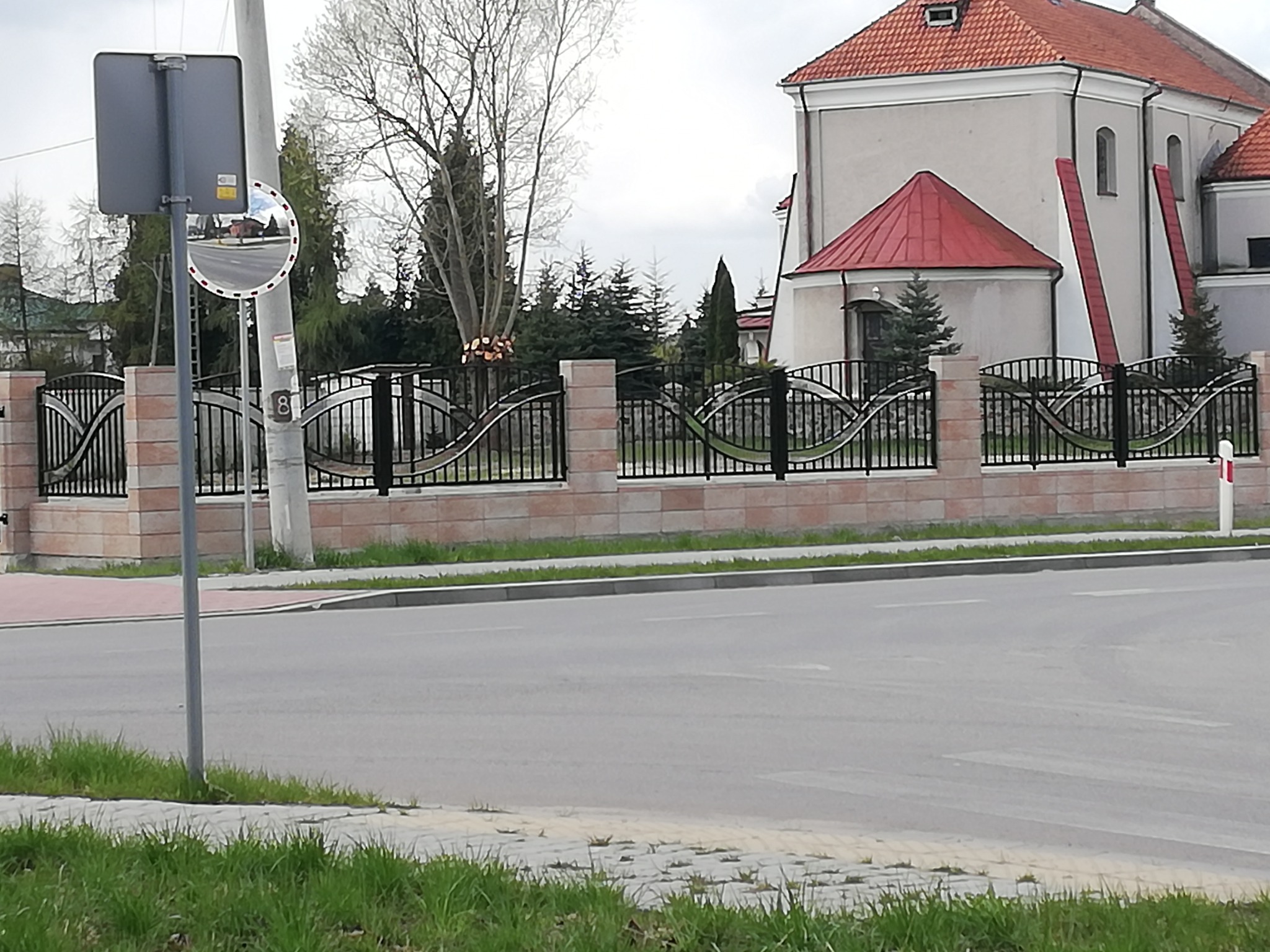 Nowoczesne ogrodzenie metalowe z falistym wzorem, osadzone na niskim murku z cegły, na tle kościoła z czerwoną dachówką i ulicy.