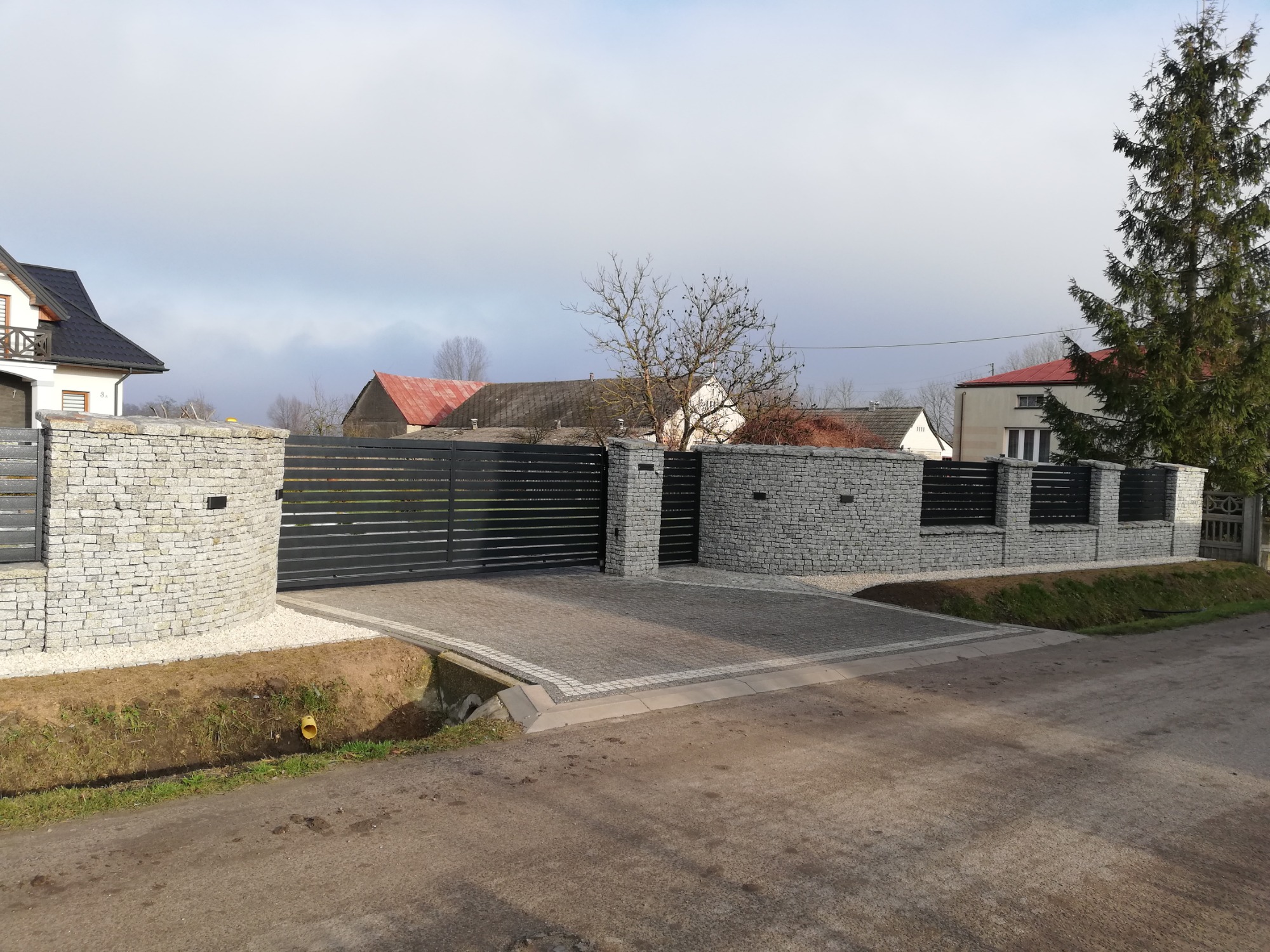 Nowoczesna posesja z przesuwną bramą i ogrodzeniem z poziomych, czarnych paneli, osadzonych na kamiennych słupkach z jasnego kamienia, wykończona jasnym żwirem, z kostką brukową na podjeździe.