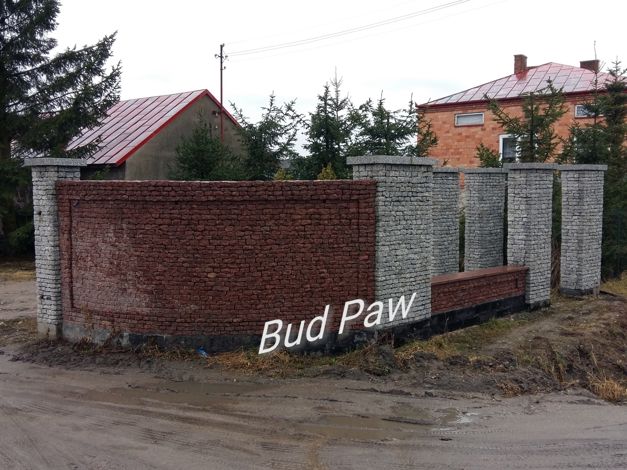 Fragment ogrodzenia z cegły klinkierowej i kamienia naturalnego, z widocznym logo firmy 'Bud Paw', na tle domów i drzew iglastych, w pochmurny dzień po deszczu.