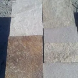 Różnokolorowe, kwadratowe płyty kamienne ułożone obok siebie, widoczne drobne nierówności powierzchni i naturalne wzory kamienia, w tle szary, sypki materiał.