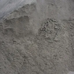 Duża pryzma szarego, drobnego materiału sypkiego z widocznymi grudkami na zboczu, ułożona pod kątem, z drzewami w tle.