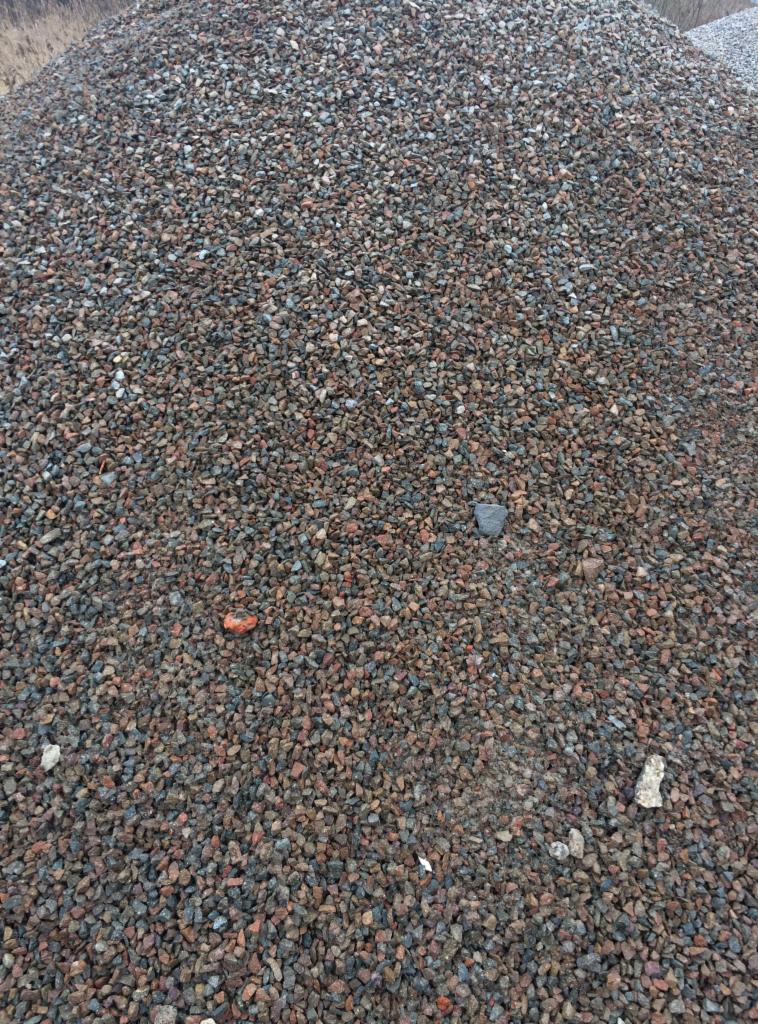 Kupa żwiru o zróżnicowanej kolorystyce, od szarego po rdzawy, z pojedynczym ceglastym elementem na pierwszym planie.