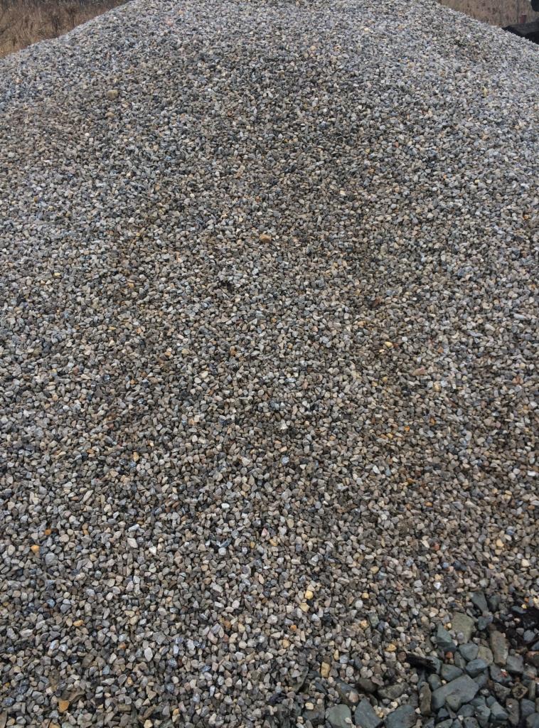 Duża pryzma drobnych, szarych kamyczków o zróżnicowanej fakturze i odcieniach, częściowo oświetlona słońcem, z widocznym fragmentem drzewa w tle.