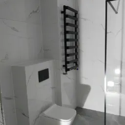 Nowoczesna łazienka z białymi płytkami imitującymi marmur, podwieszana toaleta z czarnym przyciskiem, czarny grzejnik drabinkowy i fragment szklanej kabiny prysznicowej.